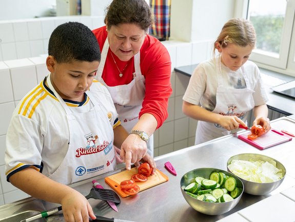 Eine Frau hilft einem Jungen und einem Mädchen beim Schneiden von Tomaten. Im Vordergrund steht eine Schüssel mit geschnittener Gurke und Salat.