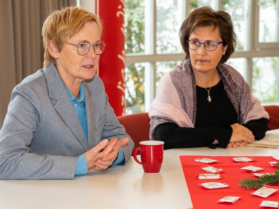 Zwei Frauen sitzen einem Tisch. Vor der linken Frau steht ein roter Becher. Im Hintergrund sind Fenster.